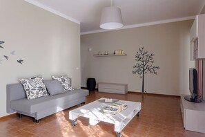 Ein Wohnzimmer mit grauem Sofa, einem weißen Couchtisch, einer Wandsticker-Darstellung eines Baumes und einem Fernseher auf einem Lowboard.