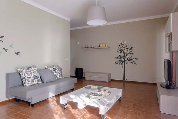 Una sala de estar con un sofá gris, una mesa de centro blanca, un adhesivo de árbol en la pared y un televisor sobre un mueble bajo.
