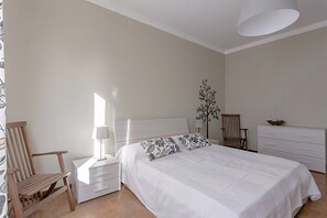 Un dormitorio con cama, dos mesitas de noche, una silla de madera, un tocador y una lámpara de pared.