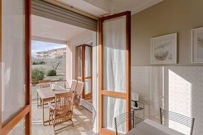 En balkon med et træbord og stole, en kaffemaskine samt udsigt til bygninger og grønne områder.