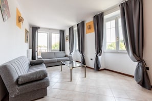 Una sala moderna con un sofá gris, una mesa de centro de vidrio y ventanas amplias con cortinas grises.