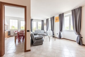 Amplia sala de estar con un sofá gris, una mesa de comedor con sillas y ventanas grandes con cortinas grises.