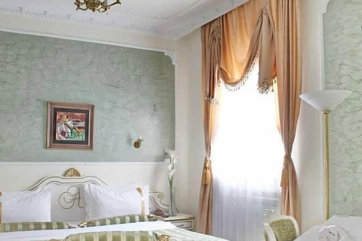 A hotel room with a bed, a desk, a chair, a chandelier, and a window with curtains.