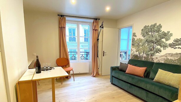 Ein Wohnzimmer mit einem grünen Sofa, einem Holztisch und einem Stuhl. Ein Fenster mit Vorhängen und eine Tür mit Pflanzenmotiv sind zu sehen.
