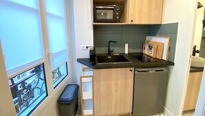 Eine kompakte Küche mit Mikrowelle, Spüle und kleinem Kühlschrank. Ein Fenster bietet Blick auf die Straße, und ein Mülleimer ist sichtbar.