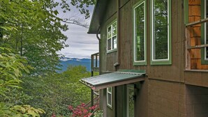 Ein Holzhaus mit Balkon, umgeben von üppiger Vegetation und Bergen im Hintergrund.