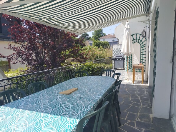 An outdoor patio with a table and chairs, a striped canopy, a grill, and a view of neighboring houses. - Séjour Vintage, Impasse sur mer (Saint-Nazaire)