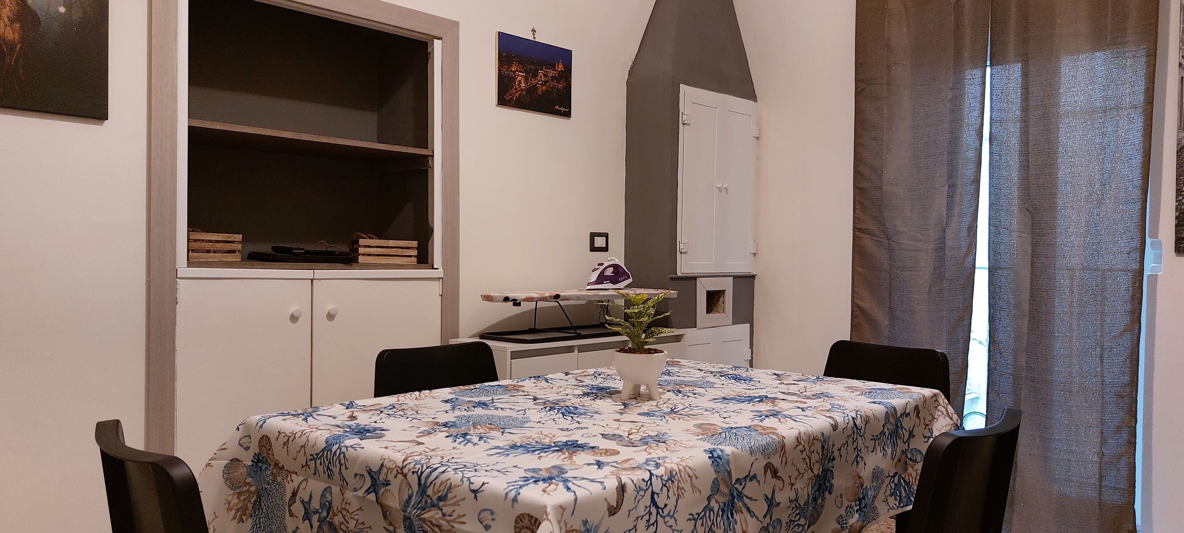 Uma área de refeições com uma mesa coberta por uma toalha florida, cercada por cadeiras. Há uma planta em vaso sobre a mesa, um armário branco com um quadro na parede e uma janela com cortinas.