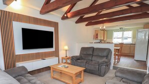 Una sala de estar con televisor de pantalla plana, pared de paneles de madera, sofás grises, una mesa de centro de madera y una cocina al fondo.