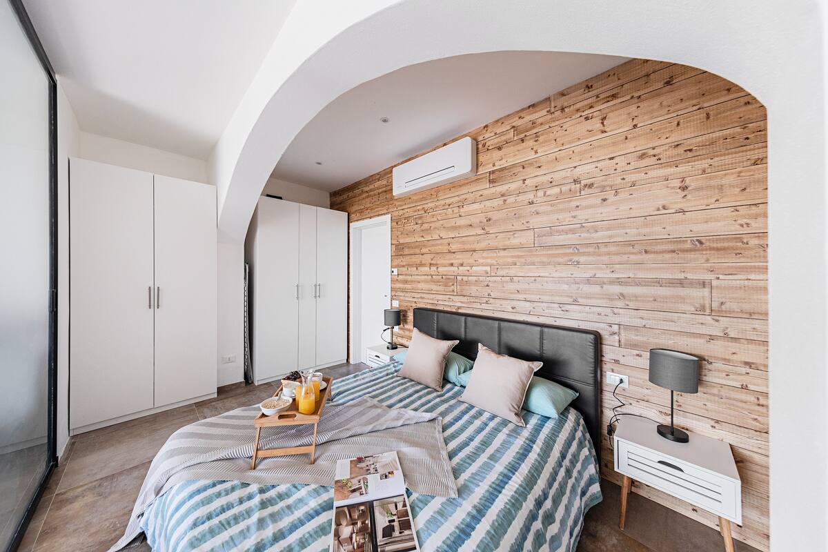 A bedroom with a wooden wall, a bed with a striped bedspread, a bedside table with a lamp, and a wardrobe.