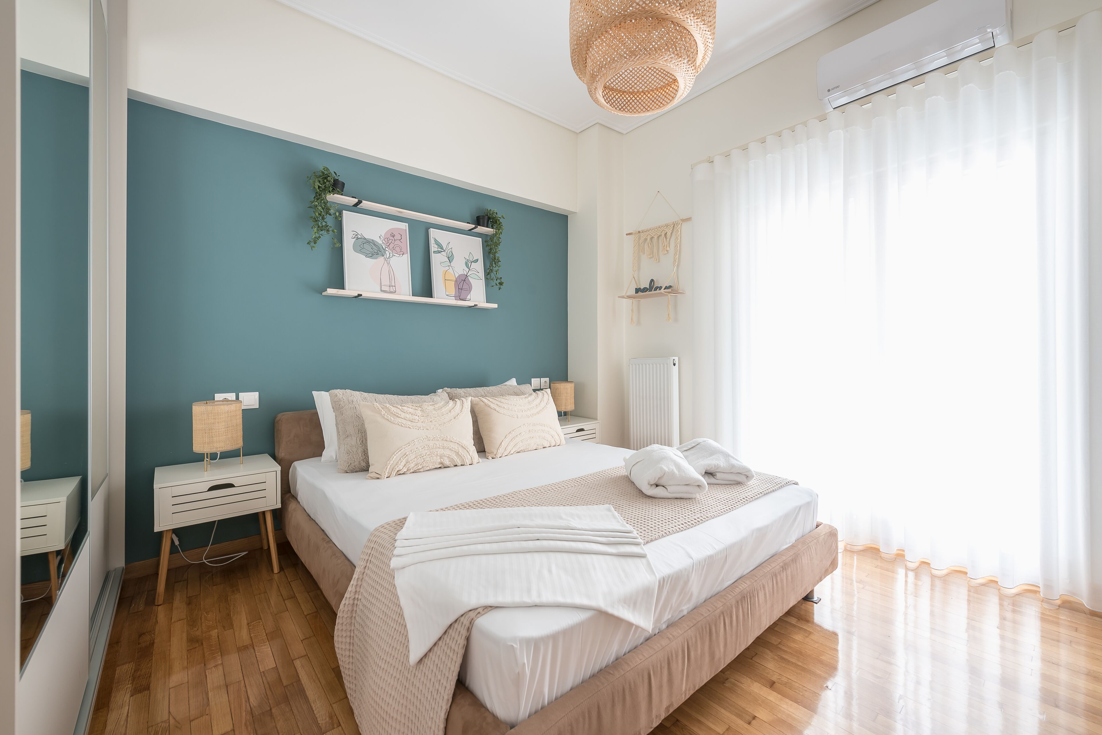 Un dormitorio con cama, mesitas de noche, un estante con obras de arte, una planta colgante y una ventana con cortinas.