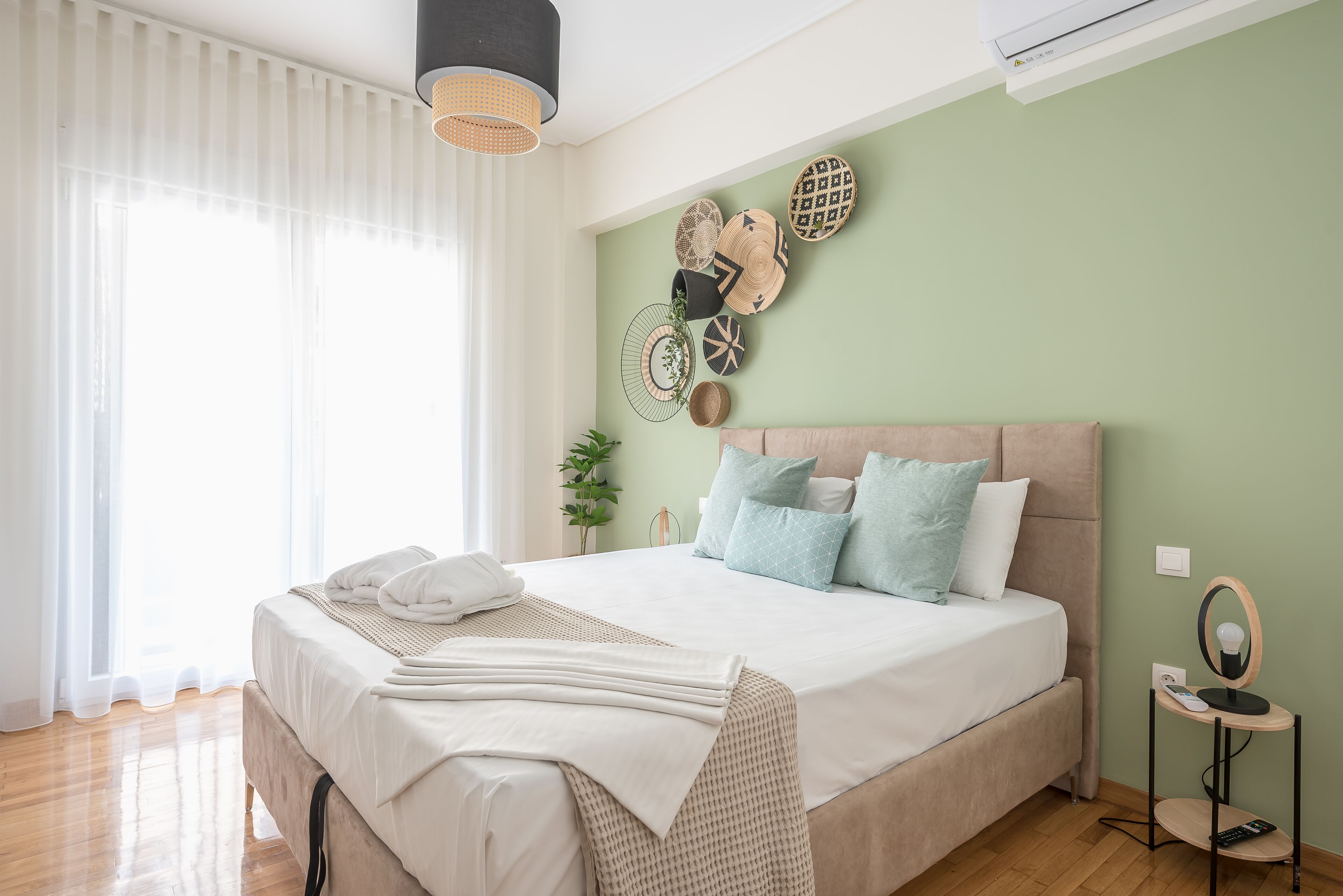 A neatly made bed with a white and beige duvet, a bedside table with a lamp, and a wall decorated with various woven baskets.