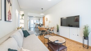 A modern living room with a sofa, coffee table, and TV. There is a dining area in the background with chairs and a table. - The Luxury Suite by Cloudkeys (Athens)