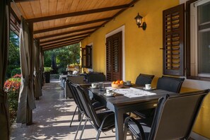A covered outdoor dining area with a table set for two, surrounded by chairs, and a yellow wall with wooden shutters. - Jolanda in Plomin  - Haus für 4-6 Personen (Plomin Luka)