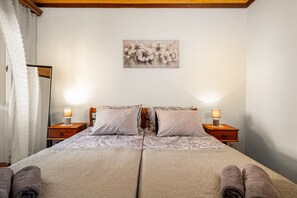 Ein Doppelbett mit gemustertem Überwurf, zwei Nachttischlampen, ein floral gestaltetes Kunstwerk an der Wand und eine Holzbalkendecke.