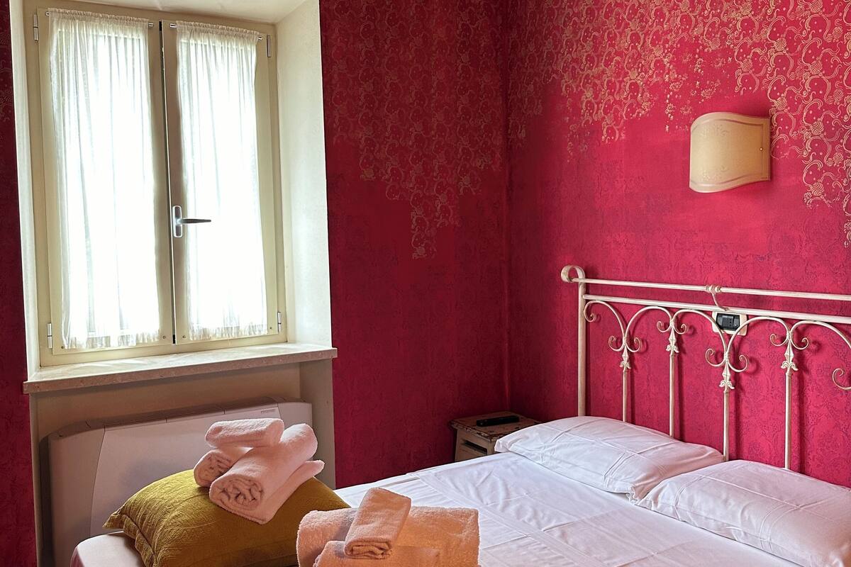 Ein Schlafzimmer mit Bett, Kissen, einem Fenster mit Vorhängen, einem Nachttisch und einer Wandleuchte.