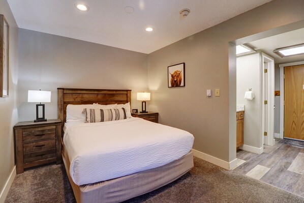 A bedroom with a large bed, two bedside tables with lamps, a wooden headboard, a framed picture on the wall, and a door leading to another room.