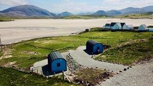 A rural landscape with a gravel path leading to blue buildings, a sandy beach, and distant mountains. - Heb Pods - North Pod
crowlista, Uig (Uig)