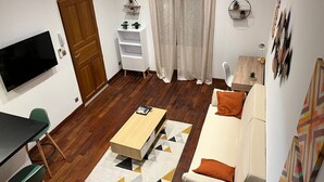 Un salon moderne avec un téléviseur à écran plat, une table basse en bois, un canapé agrémenté d’un coussin orange et une étagère blanche ornée de plantes.