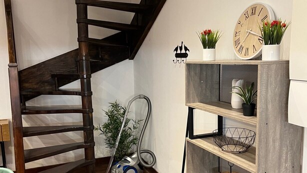 Un escalier en colimaçon en bois, un plancher en bois, une bibliothèque agrémentée d’objets décoratifs et une horloge au mur.