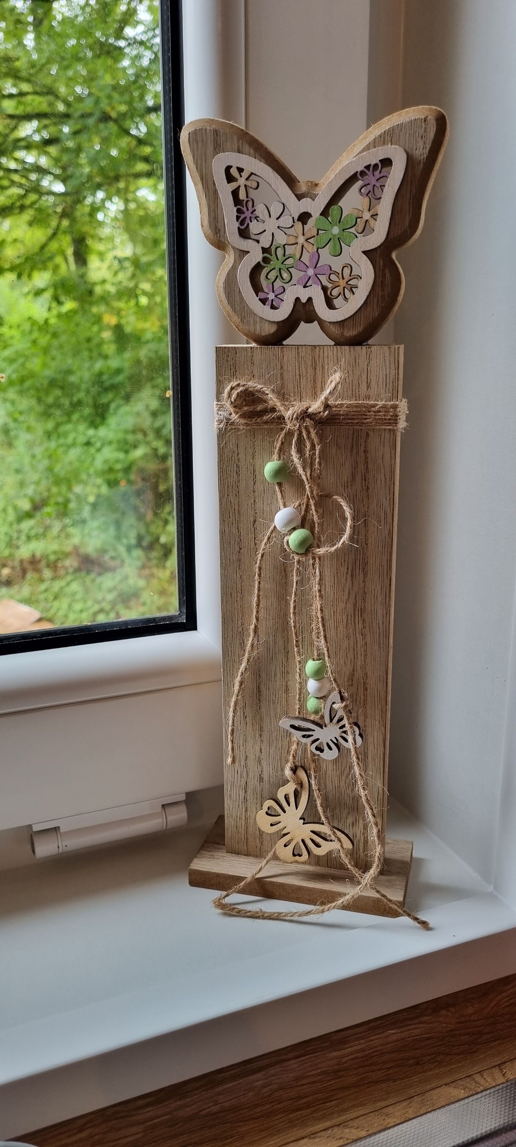 Ein hölzernes Dekorationsobjekt mit Schmetterlings- und Blumenmuster, verbunden mit Juteband und Perlen, auf einem Fensterbrett platziert.