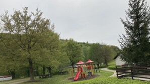 Leikkikenttä, jossa on punainen liukumäki ja keltaisia rakenteita, ja sen ympärillä on puita ja ruohikkoista aluetta.