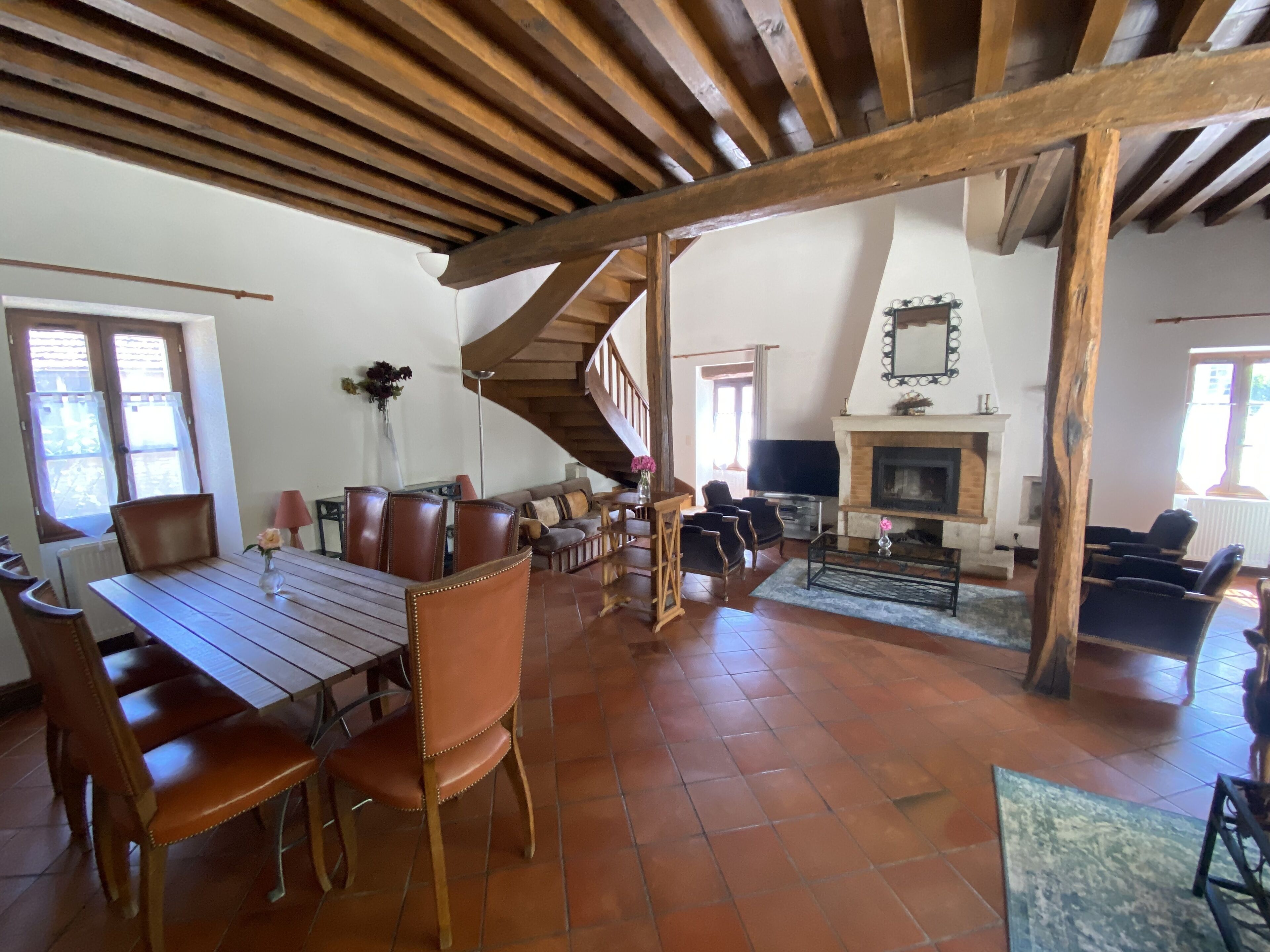 A spacious living area with a wooden ceiling, a fireplace, a dining table with chairs, and a staircase.