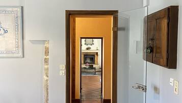A hallway with a tiled floor, wooden beams on the ceiling, a door leading to a room with a fireplace, and a wall-mounted picture.