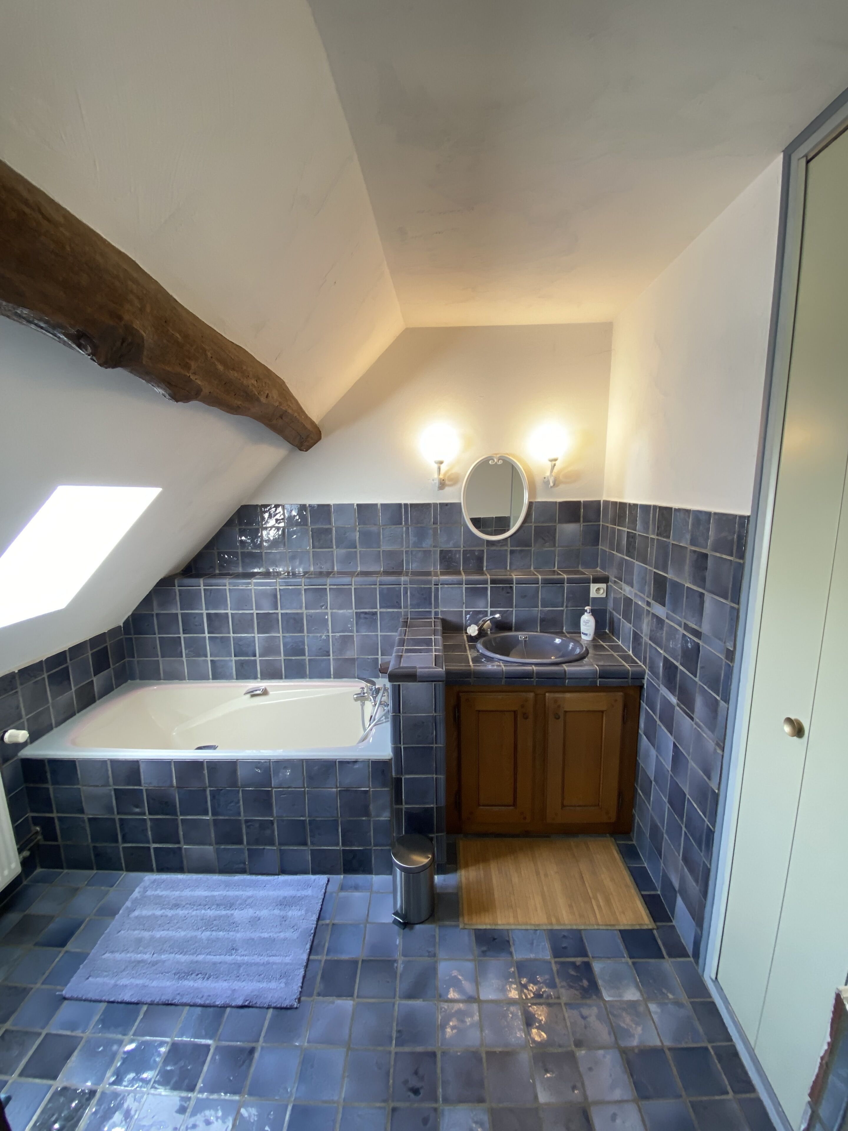 Un baño con bañera, lavamanos y espejo. Hay un tragaluz sobre la bañera y las paredes están revestidas con azulejos.