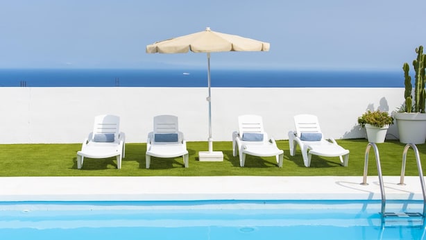 Un espace aménagé au bord de la piscine, avec des chaises longues blanches et un parasol beige, offrant une vue sur un ciel bleu azur et l’océan.