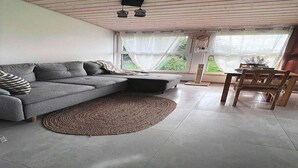 A modern living room with a grey sectional sofa, a round woven rug, a wooden dining table with chairs, and a large window with curtains. - Holiday Home near Baltic Sea & Golf Course (Kołczewo)