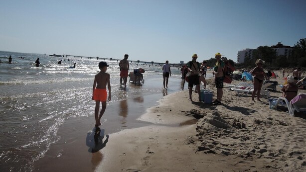 Una spiaggia con persone che camminano sulla riva, alcune in acqua, e sedie a sdraio e ombrelloni sulla sabbia.