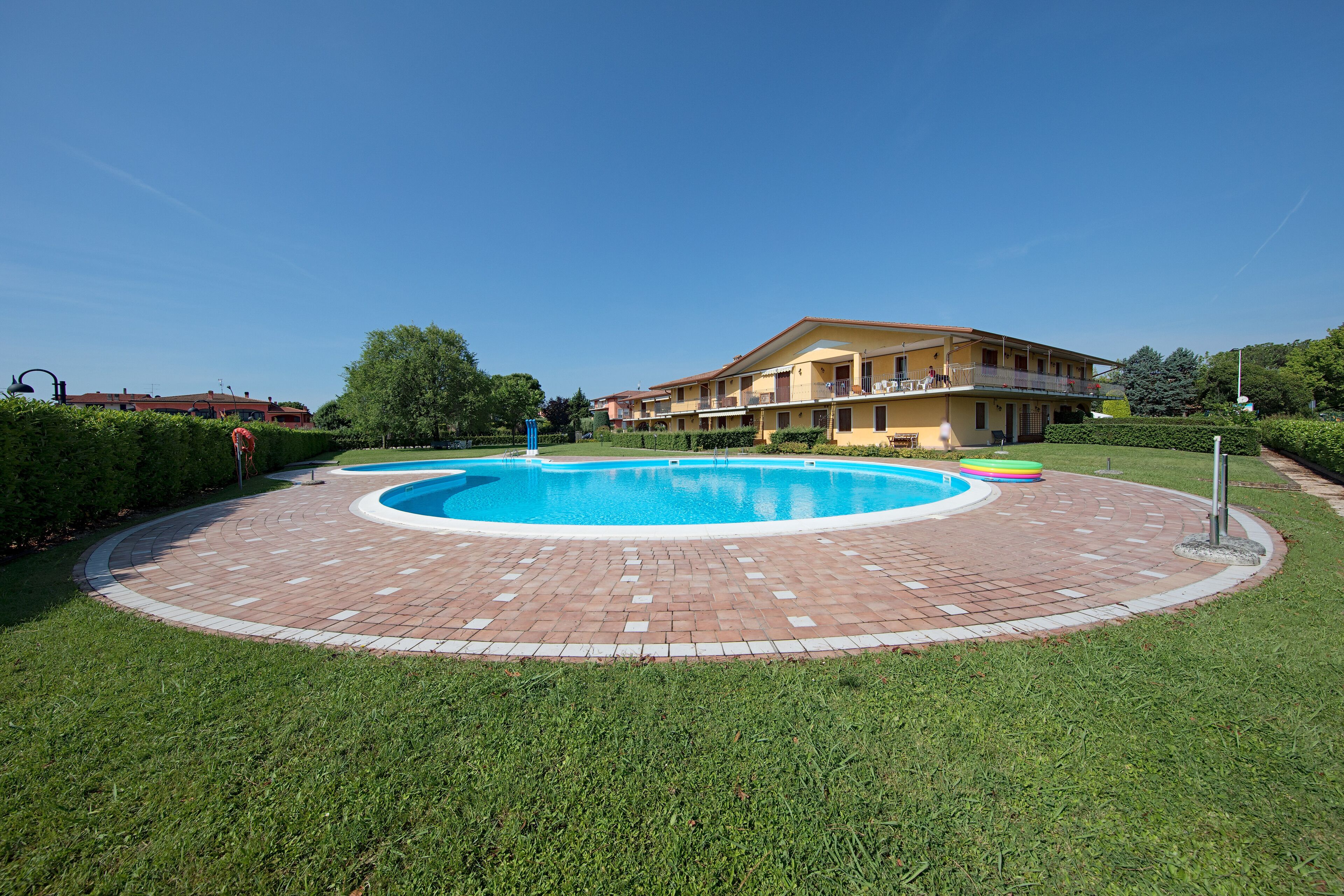 A circular pool with a brick border, surrounded by well-maintained grass, and a building with balconies in the background.