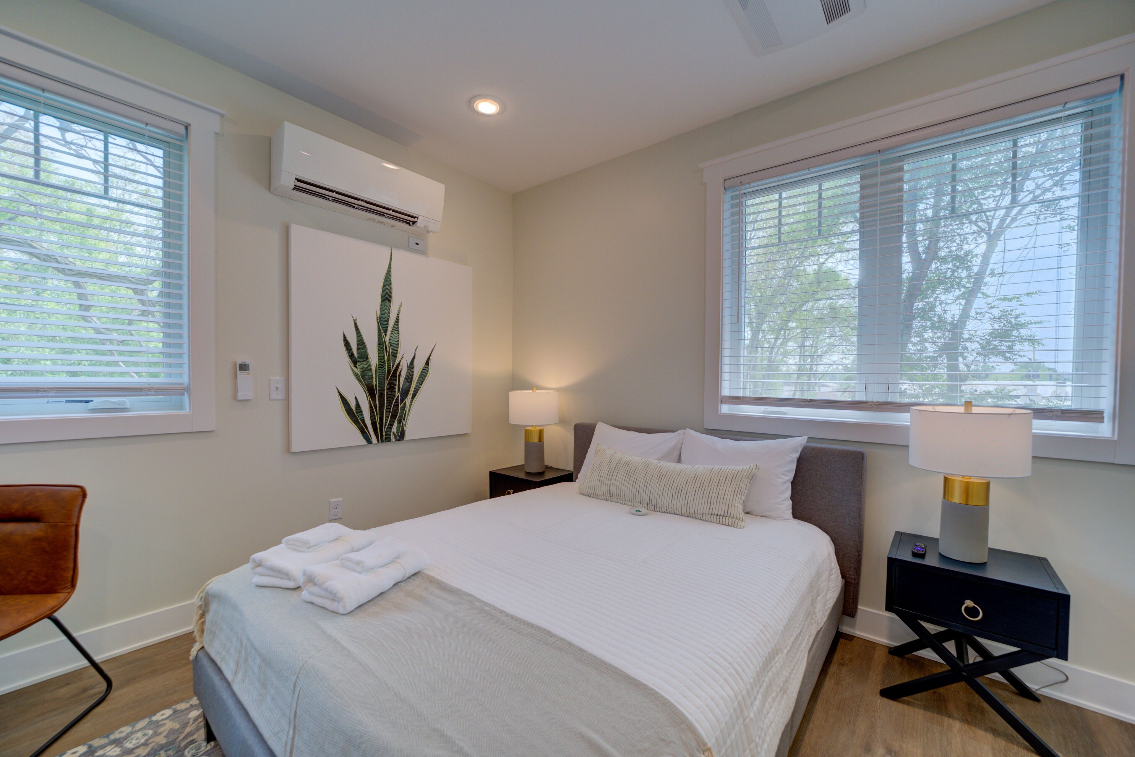 A modern bedroom with a large bed, two bedside tables with lamps, a wall-mounted air conditioner, and a window with blinds.