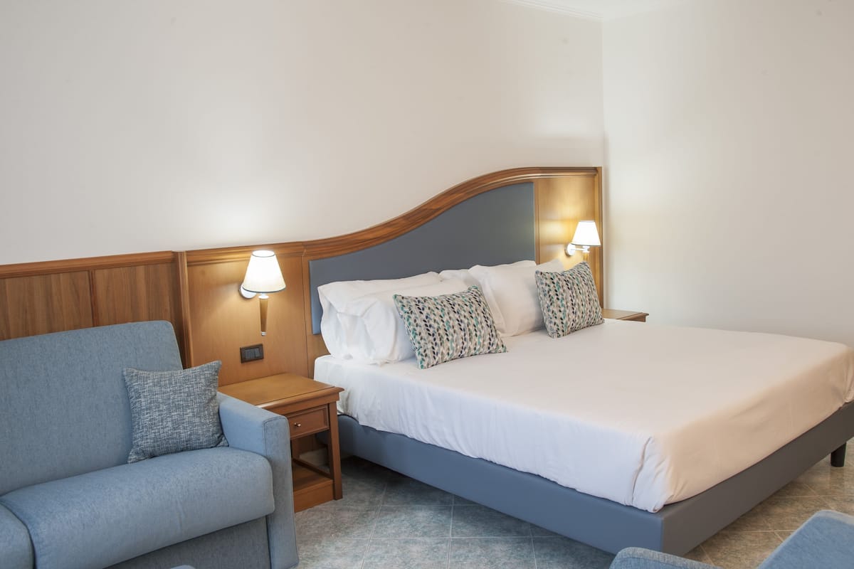 A bedroom with a bed, a nightstand, a lamp, a sofa, and a patterned pillow.