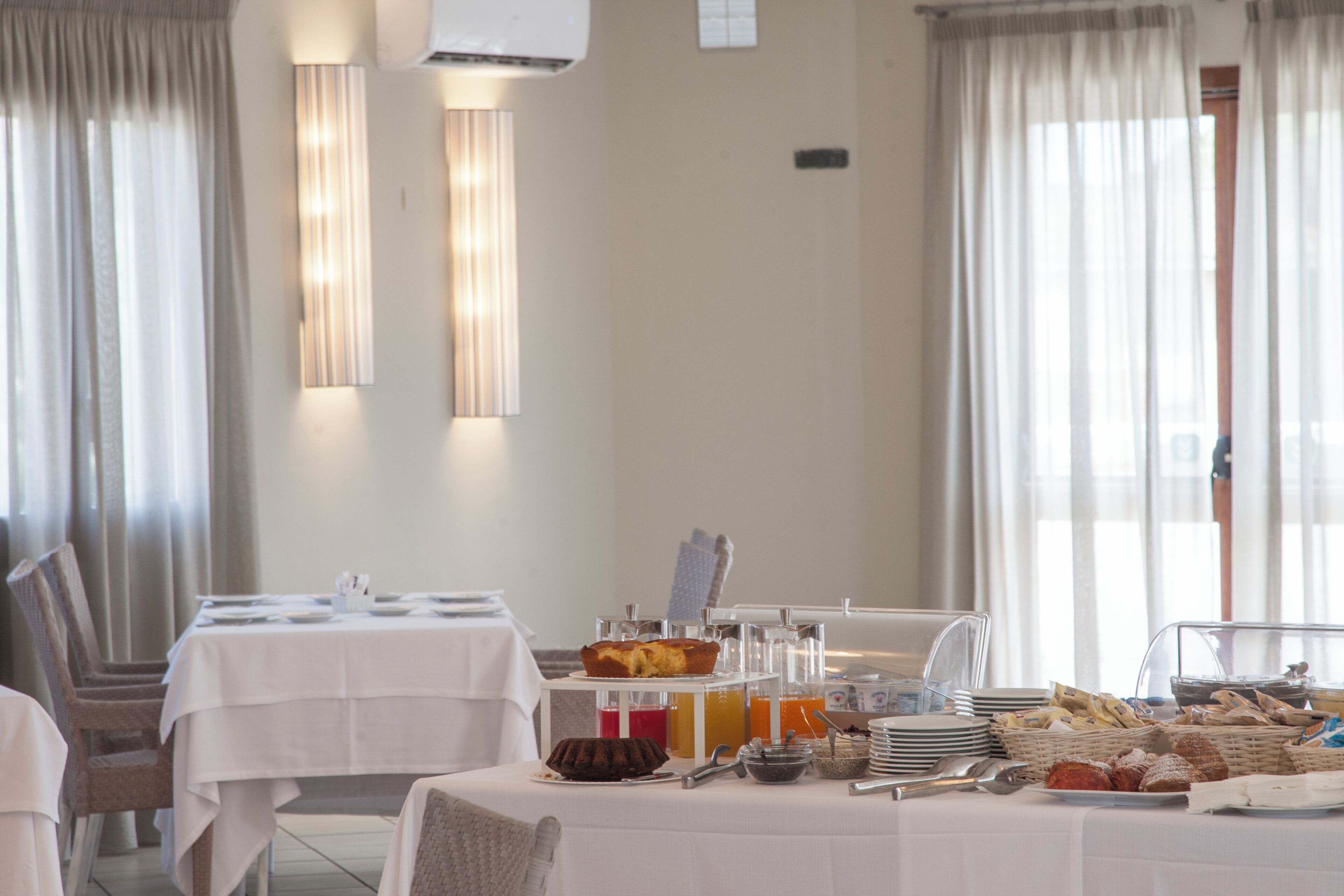 Un buffet de petit-déjeuner est installé sur une table, avec divers aliments et ustensiles, et est entouré de chaises et de rideaux.