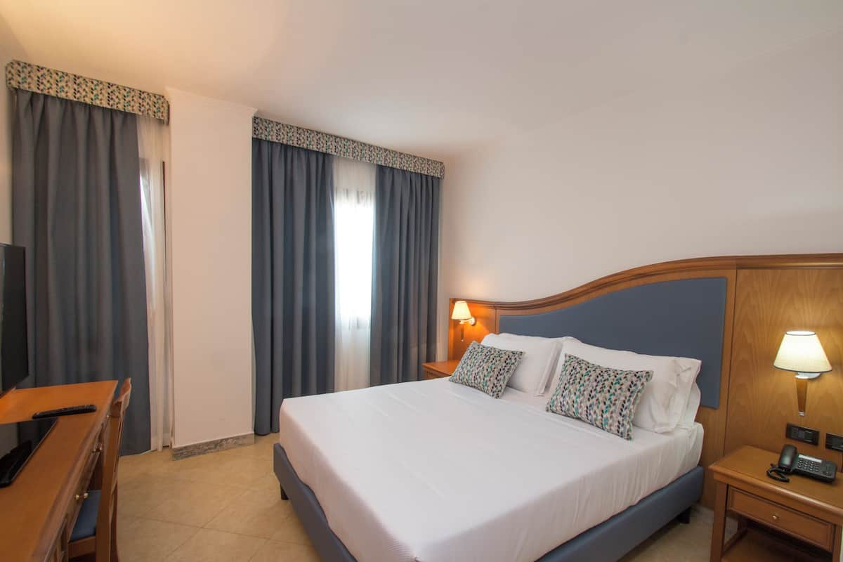 A hotel room with a large bed, a desk, a chair, a bedside table with a lamp, and a TV.