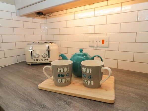A kitchen with white tiled walls, a wooden countertop, a white toaster, a teal teapot, and two mugs with the text "MAKE me a CUPPA".