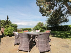 An outdoor patio with wicker furniture and a glass table, surrounded by greenery and a clear sky.