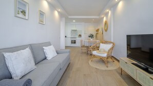 Een moderne woonkamer met een grijze bank, een rieten stoel, een televisie en een eethoek met een tafel en stoelen.