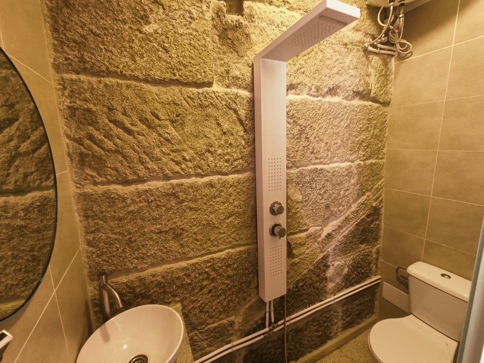 A bathroom with a stone wall, a white sink, a toilet, and a shower with a control panel.