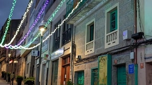Una calle bordeada de edificios, algunos con balcones y contraventanas verdes, iluminada por luces colgantes.