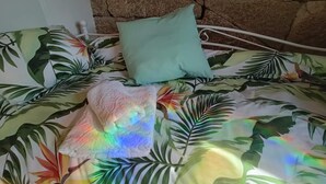 Una cama con colcha de estampado de hojas tropicales, un cojín azul claro y una toalla de colores del arcoíris.