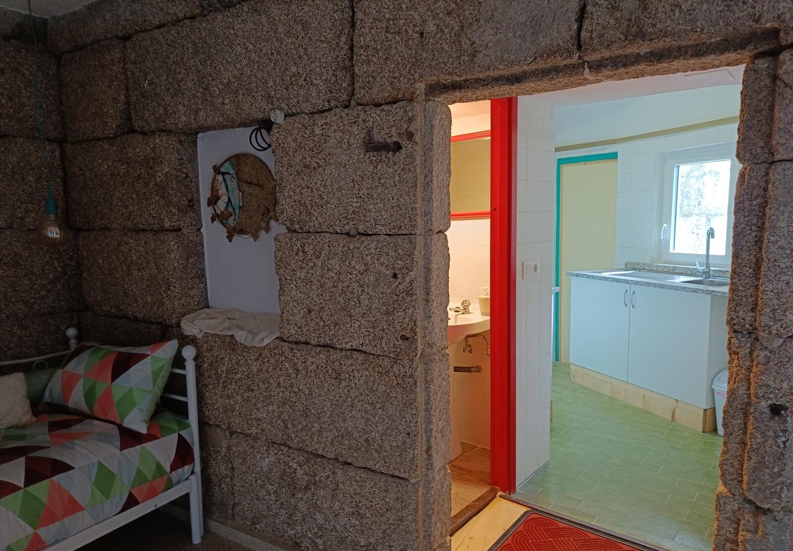 A room with a bed, a stone wall, and a doorway leading to a kitchen with white cabinets and a window.