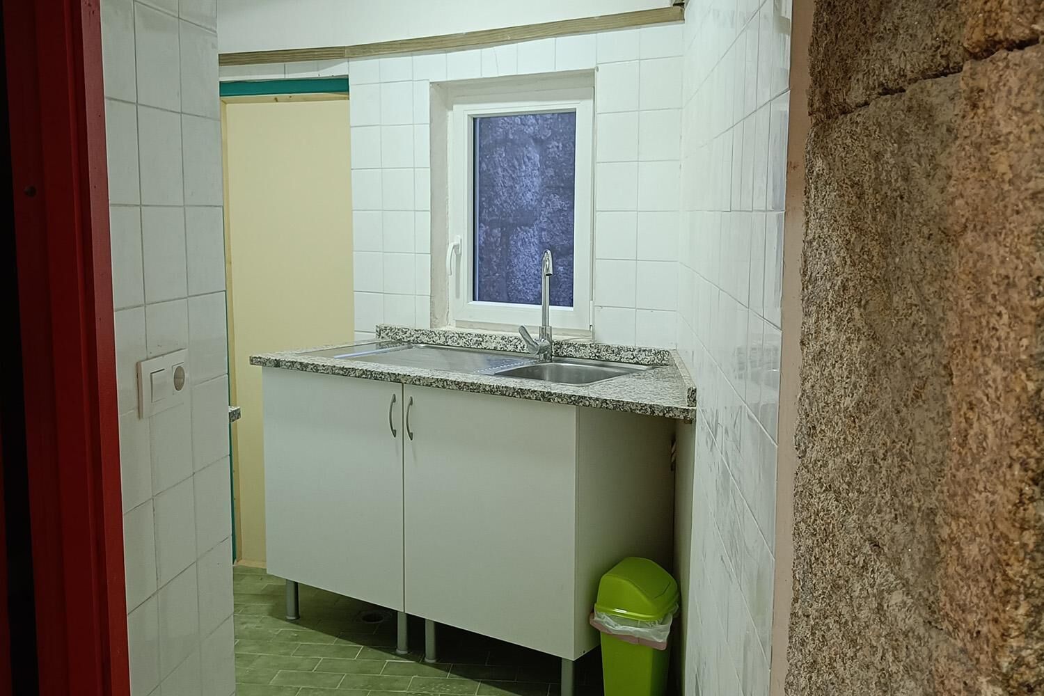 Una cocina pequeña con armarios blancos, una encimera de granito, un ventanal sobre el fregadero y un cubo de basura verde.