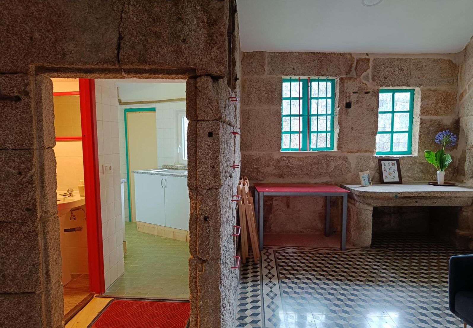 A room with a stone wall, a small table with a red top, a window with green shutters, and a tiled floor.