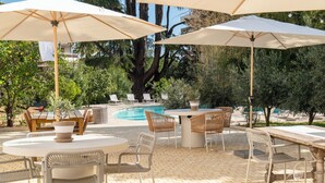 An outdoor patio with round tables and chairs, umbrellas, and a pool area surrounded by trees. - La Bâtisse en Blanc (Vallauris)