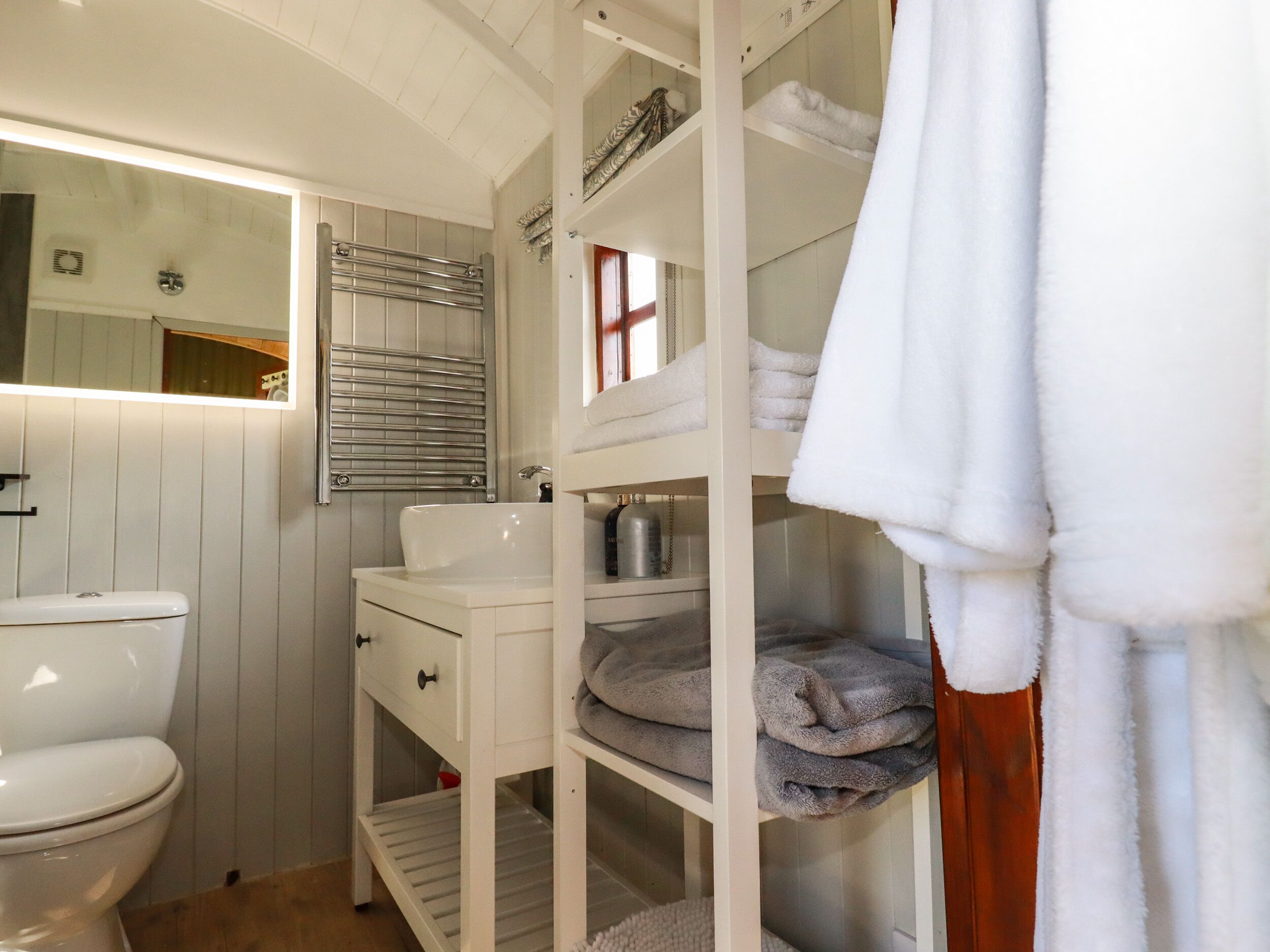 Un baño pequeño con inodoro blanco, lavamanos y espejo. Se observan repisas con toallas dobladas y artículos de tocador.