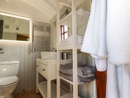Un baño pequeño con inodoro blanco, lavamanos y espejo. Se observan repisas con toallas dobladas y artículos de tocador.