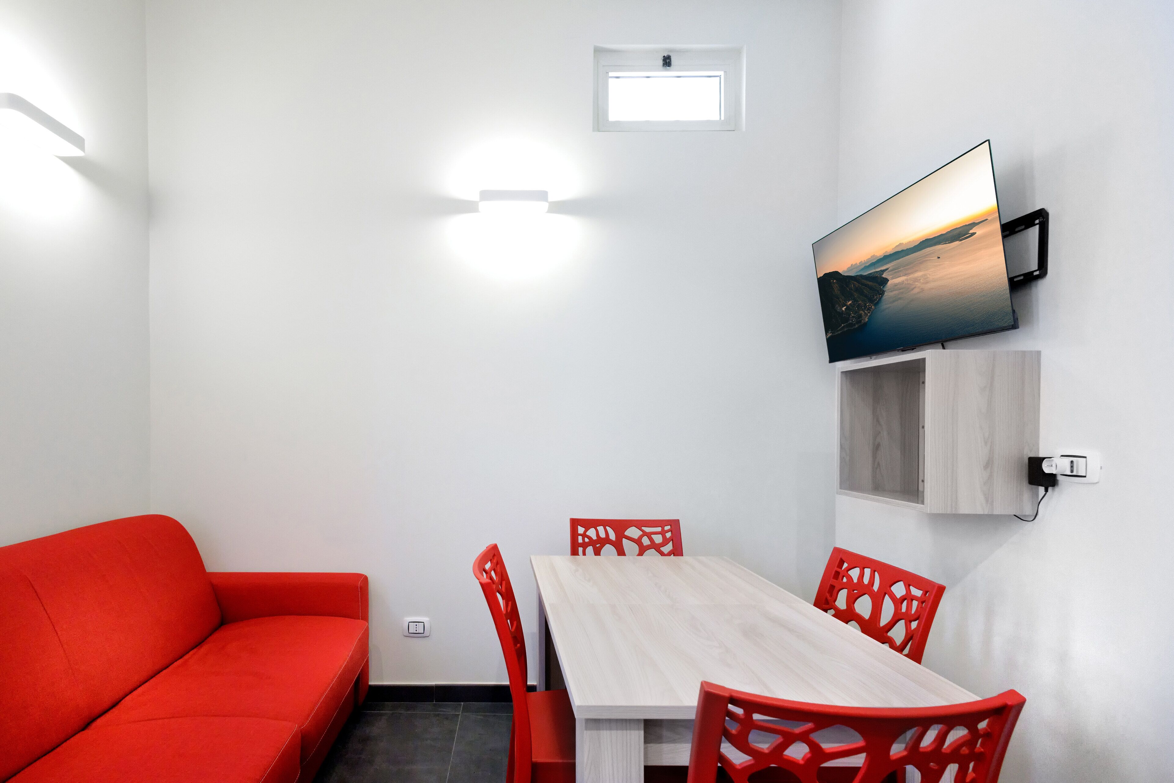Een moderne kamer met een rode bank, een houten eettafel met rode stoelen, een wandgemonteerde televisie en een klein raam.
